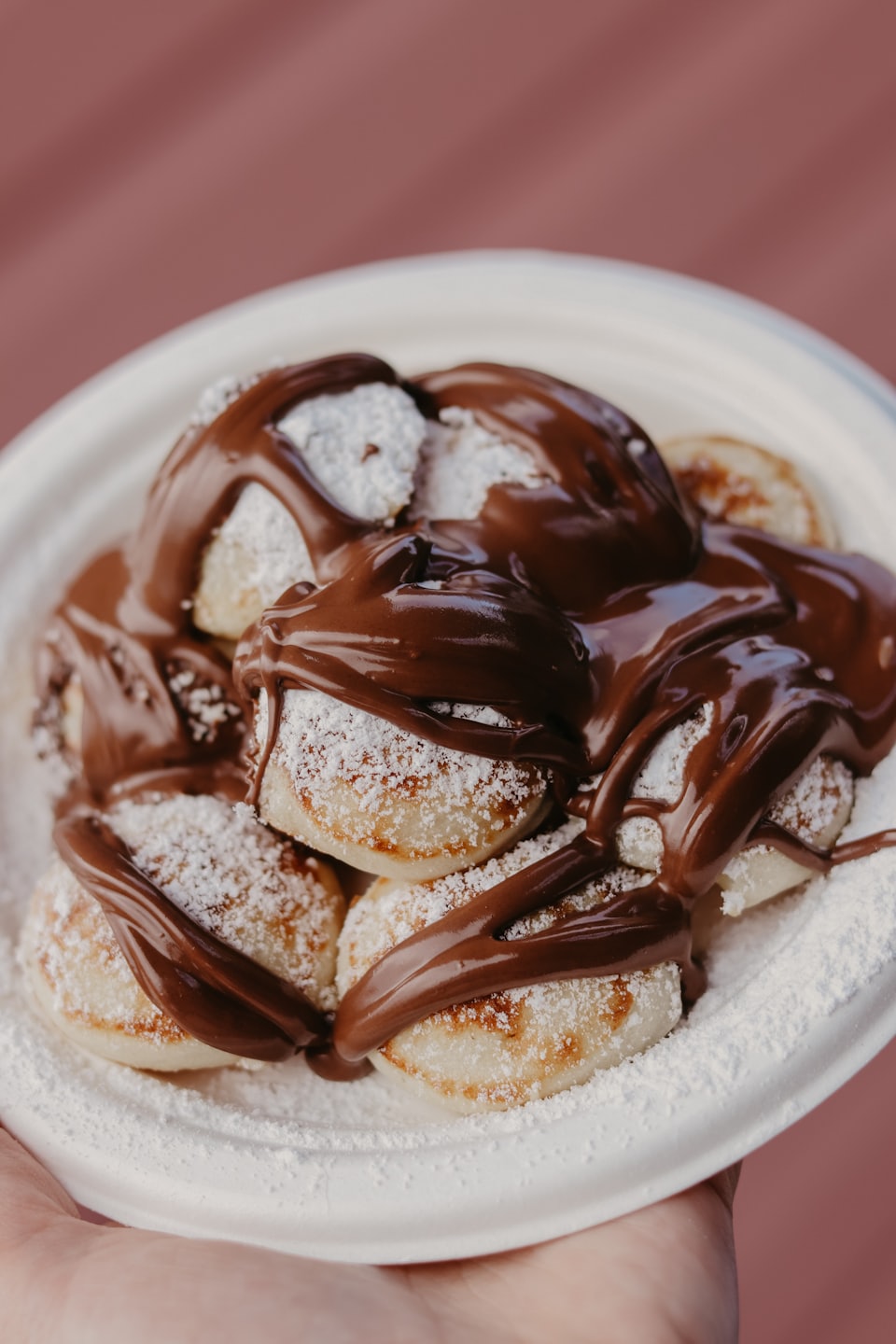 A hand holds a plate of small pancakes with chocolate sauce.